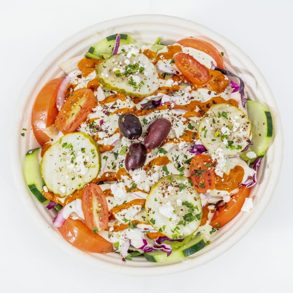 Greek salad bowl topped with cucumbers, tomatoes, olives, feta cheese, pickles, herbs, and creamy orange dressing.