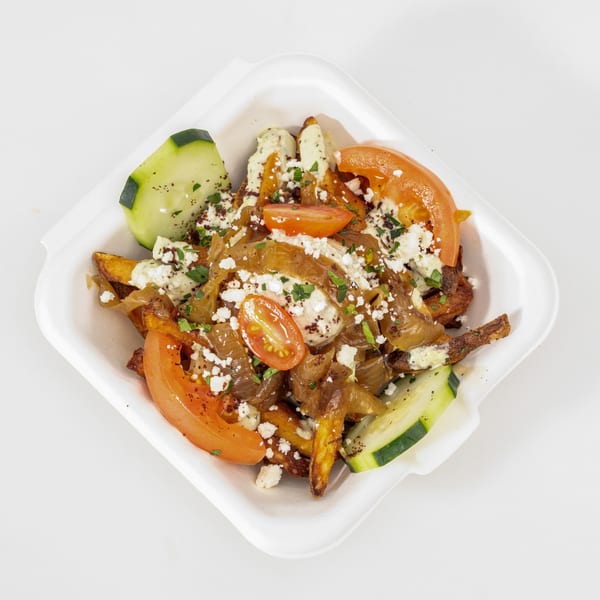 Loaded fries topped with tomatoes, cucumbers, caramelized onions, creamy sauce, crumbled cheese, and herbs in a white takeout tray.