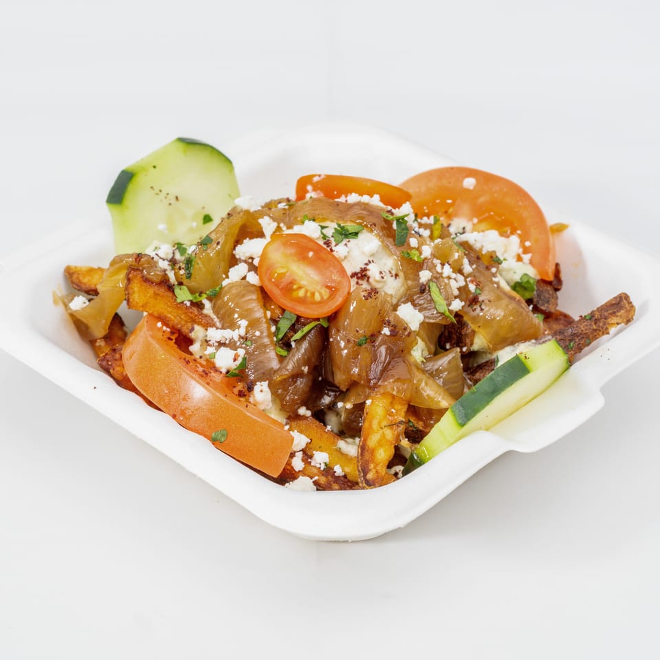Loaded fries topped with grilled onions, tomatoes, cucumber, feta cheese, herbs, and creamy sauce in a white takeout container.