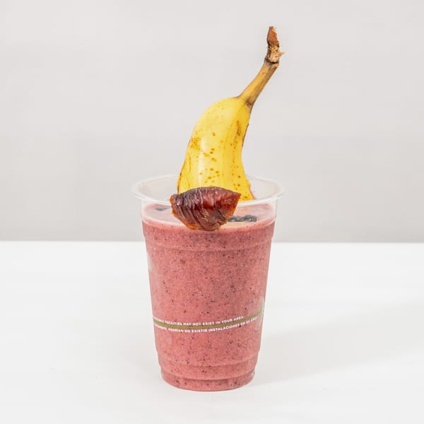 Pink berry smoothie topped with blueberries, a banana, and a dried fruit garnish in a clear cup.