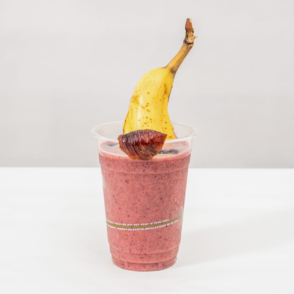 Pink berry smoothie topped with blueberries, a banana, and a dried fruit garnish in a clear cup.