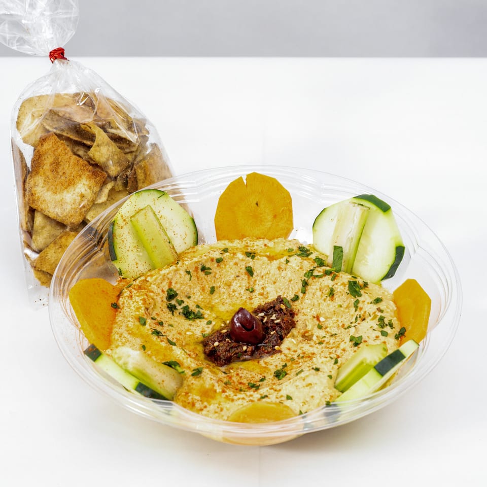 Hummus platter garnished with olives, herbs, cucumbers, and carrots, served with pita chips in a clear container.