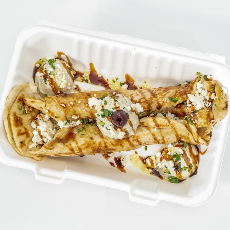 Grilled folded flatbreads with fries, feta, herb sauce, olives, and balsamic drizzle in a takeout container.